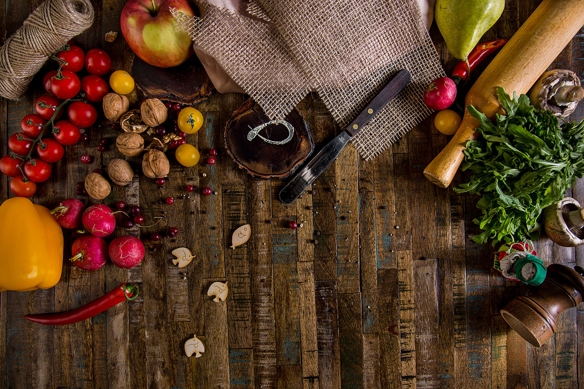 Wooden background with cherry tomatoes, apple, butter knife, rolling pin, linen cloth, linen thread, apple, paprika, walnut, chili pepper, pear, arugula, champignons and cranberries